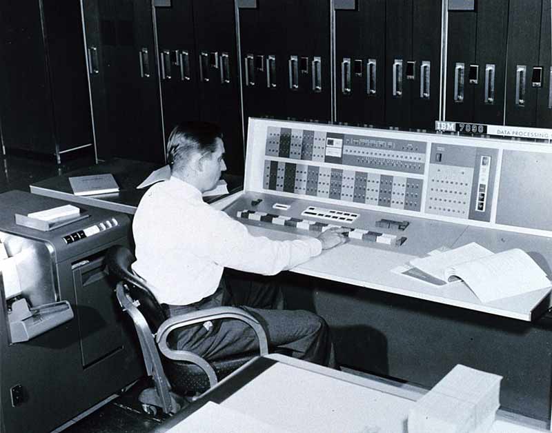 A man in a white shirt pressing a button on a 1960's IBM computer console